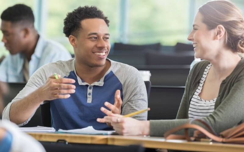 A young man smiles as he sits and talks to a female classmate at his desk