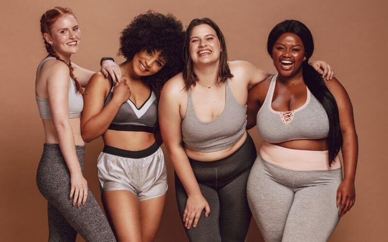 Group of women of different race, figure type and size in sportswear standing together over brown background