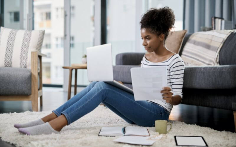 An image of a young woman sitting with paperwork while working on her laptop